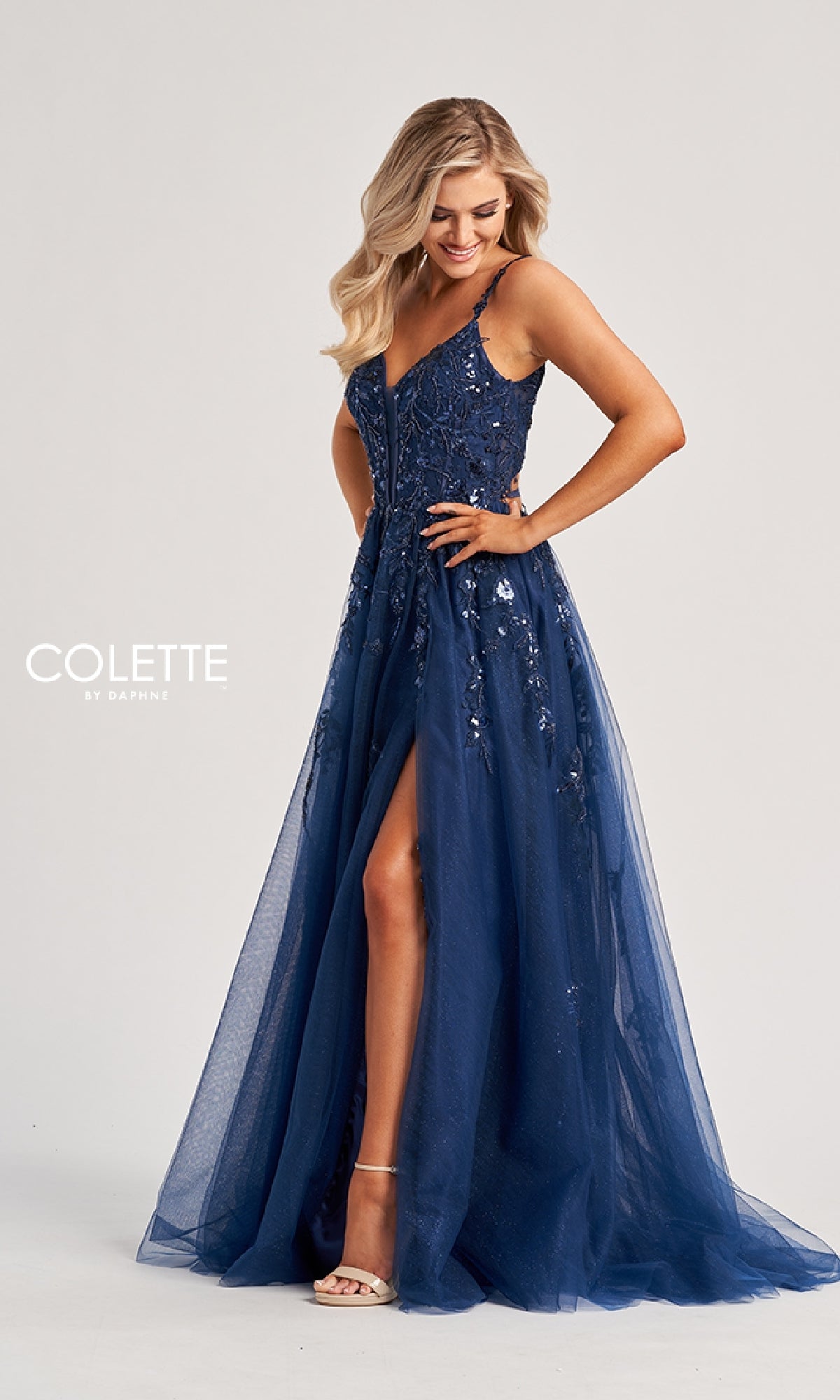 A woman wears the Colette CL8060 long prom dress—a sleeveless, floor-length navy blue sequin gown with a thigh-high slit and elegant open back—standing against a plain light background.